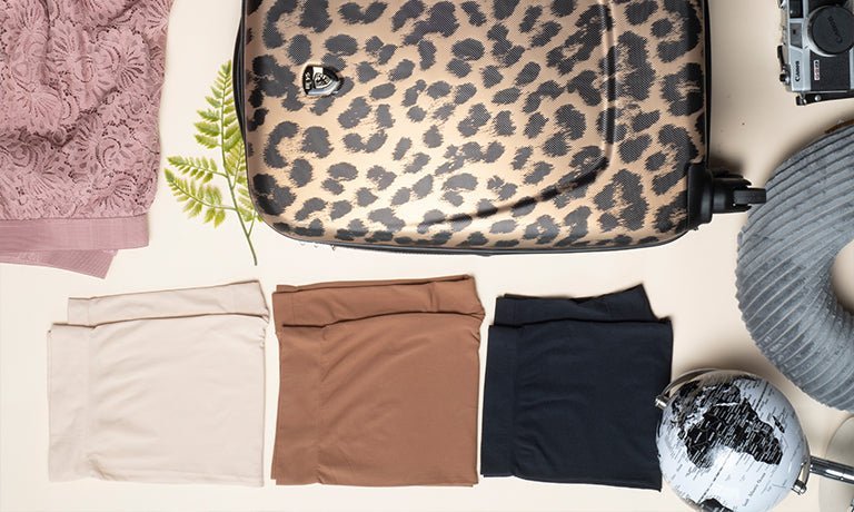 Flat lay of travel essentials: leopard-print carry-on, three folded anti-chafing shorts in nude, brown, and black, neck pillow, camera, and small globe