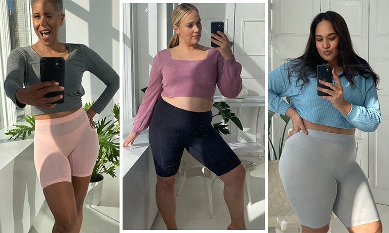 Three women taking mirror selfies in a bright studio, wearing high-waist mid-thigh biker shorts (pink, black, light gray) and cropped tops