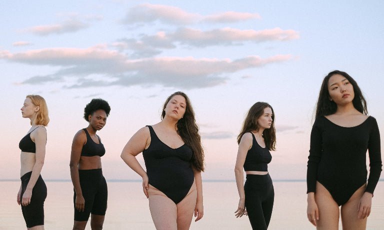 Five diverse women wearing black swimsuits and bodysuits standing on a beach at sunset
