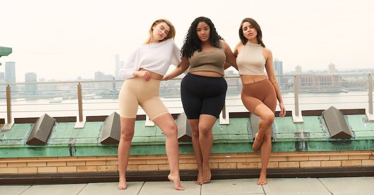 Three models wearing beige, black, and brown mid-thigh anti-chafing shorts posing on a rooftop with the city skyline behind them