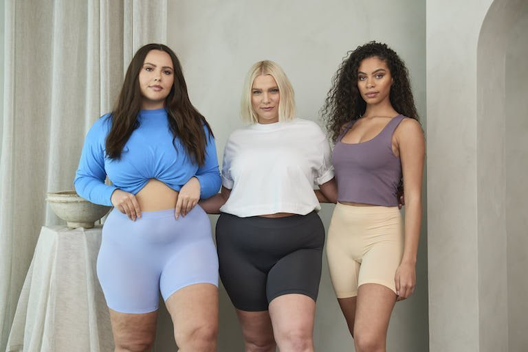 Three women modeling high-waisted biker shorts in lavender, black, and beige standing arm-in-arm against a neutral studio backdrop
