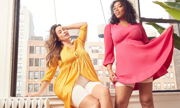 Two women by a sunlit city window, one in a yellow dress with beige mid-thigh shorts, the other in a pink dress showing beige shorts