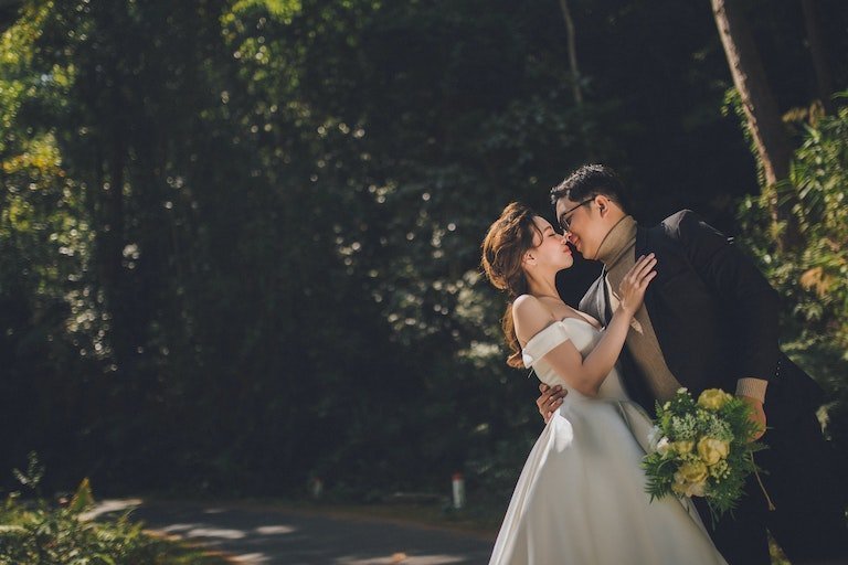 Bride in a white fitted off-shoulder gown and groom in a dark suit embracing on a shaded forest road, bride holding a yellow bouquet