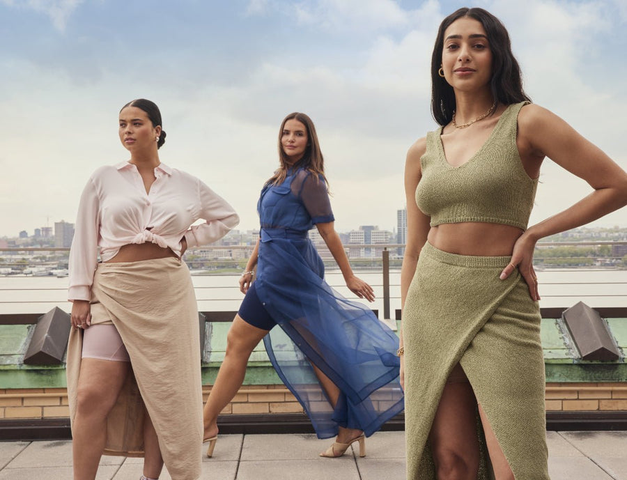 Three women posing on a rooftop with a city view: beige skirt, blue sheer dress, olive-green crop top and skirt