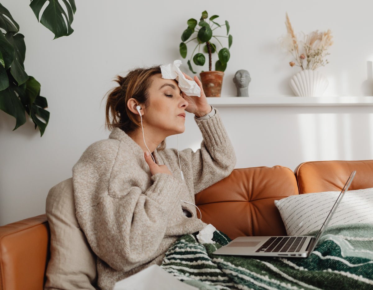 Woman holding a tissue to her forehead on a couch, wearing earbuds with a laptop nearby and houseplants on a shelf