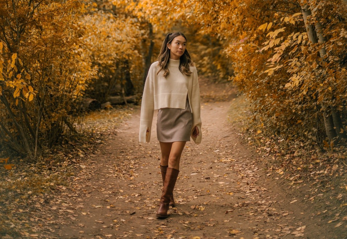 Woman walking on a leaf-covered path wearing a cream cropped sweater, plaid mini skirt, and brown knee-high boots amid autumn foliage