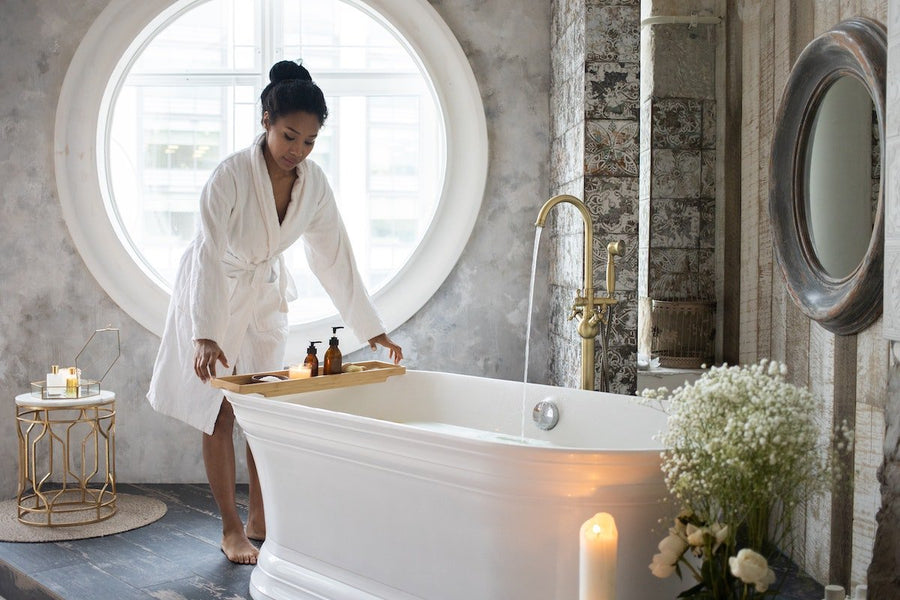 Woman in white bathrobe placing a wooden tray across a freestanding bathtub in a bright spa-like bathroom with round window and lit candles