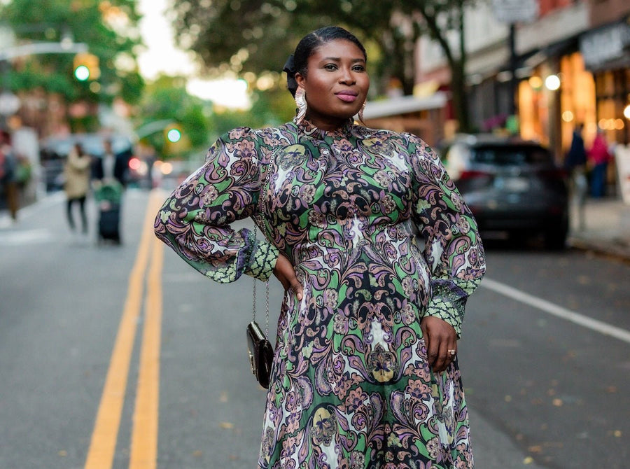 Woman wearing a long multicolor paisley dress fitted at the waist, posing on a city street at dusk