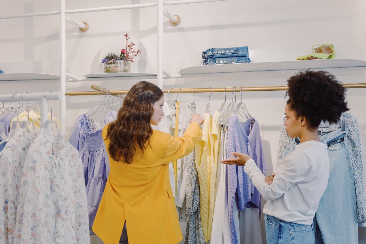 Shopper in a yellow blazer browsing pastel and denim garments on a boutique rack while an employee gestures nearby