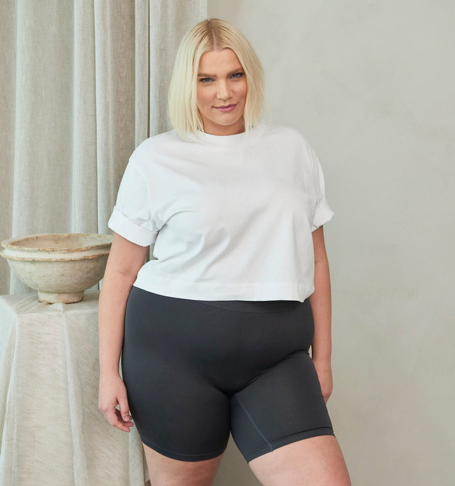 Plus-size model standing wearing mid-thigh black biker shorts and a white cropped tee in a neutral studio