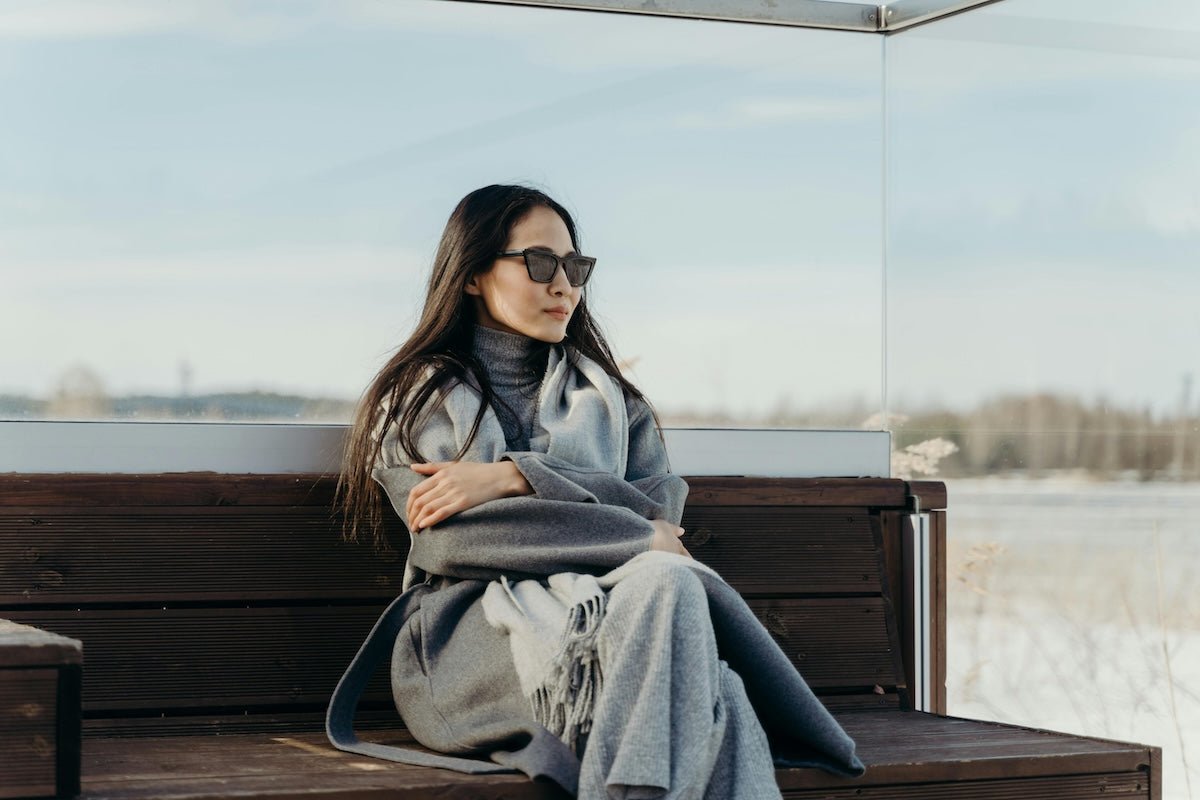 Woman wearing a gray coat, turtleneck, scarf and sunglasses sitting on a wooden bench overlooking a snowy field
