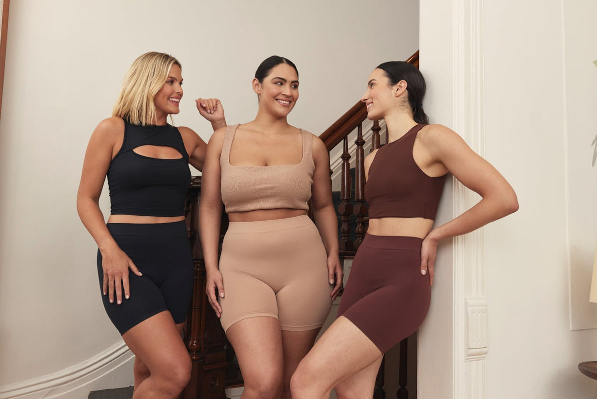 Three models smiling and chatting while wearing Thigh Society biker shorts and cropped tops in black, nude, and brown on a staircase