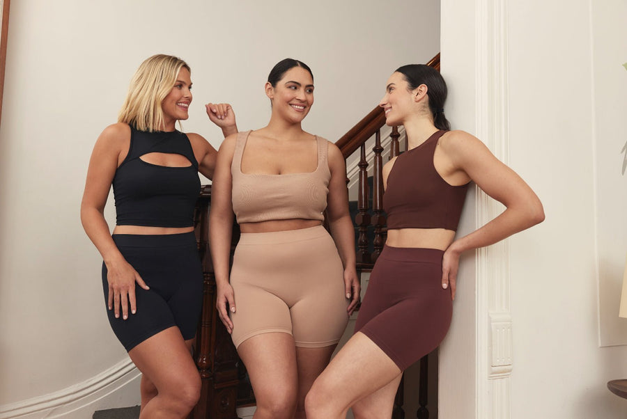 Three models smiling and chatting while wearing Thigh Society biker shorts and cropped tops in black, nude, and brown on a staircase