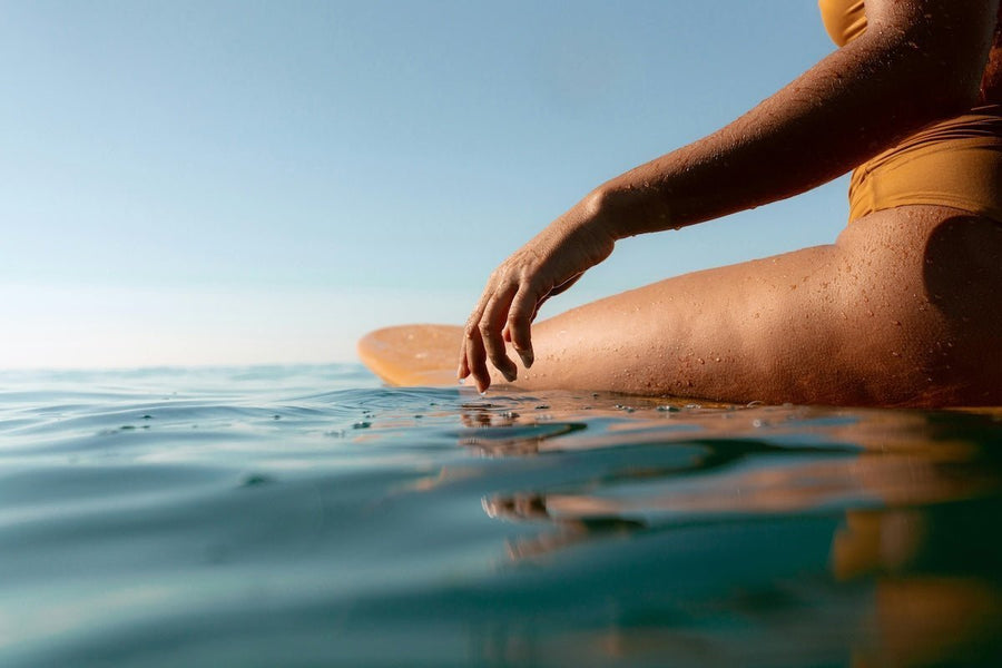 Close-up of a woman wearing a yellow swimsuit sitting on a surfboard, hand skimming calm ocean water