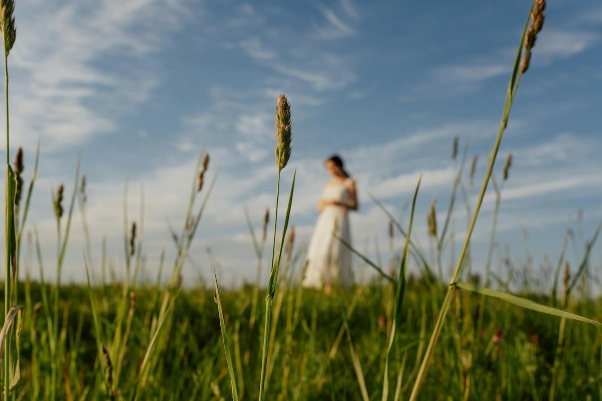 Woman standing in a white sundress in a sunlit grassy field with tall grasses in sharp focus in the foreground