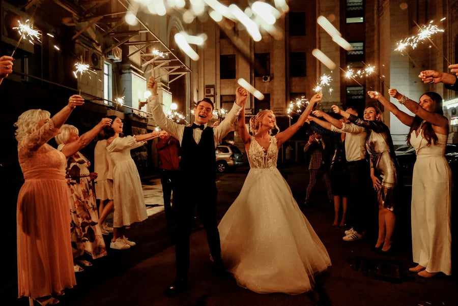 Bride in a lace wedding gown and groom in a tux celebrate as guests hold sparklers during a nighttime wedding exit on a city street