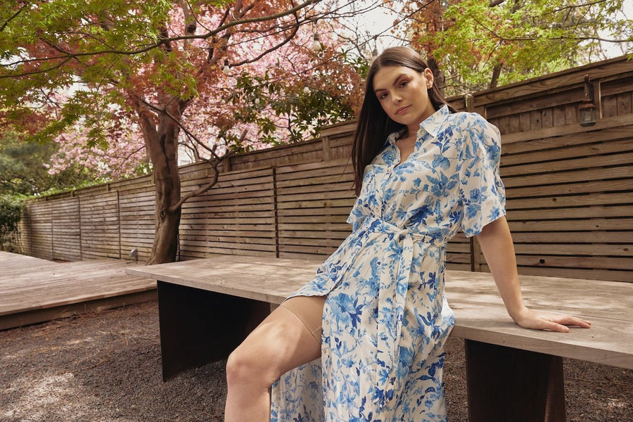 Woman seated on a wooden bench showing a beige anti chafing thigh band under a white and blue floral dress in a fenced garden with pink blossoms
