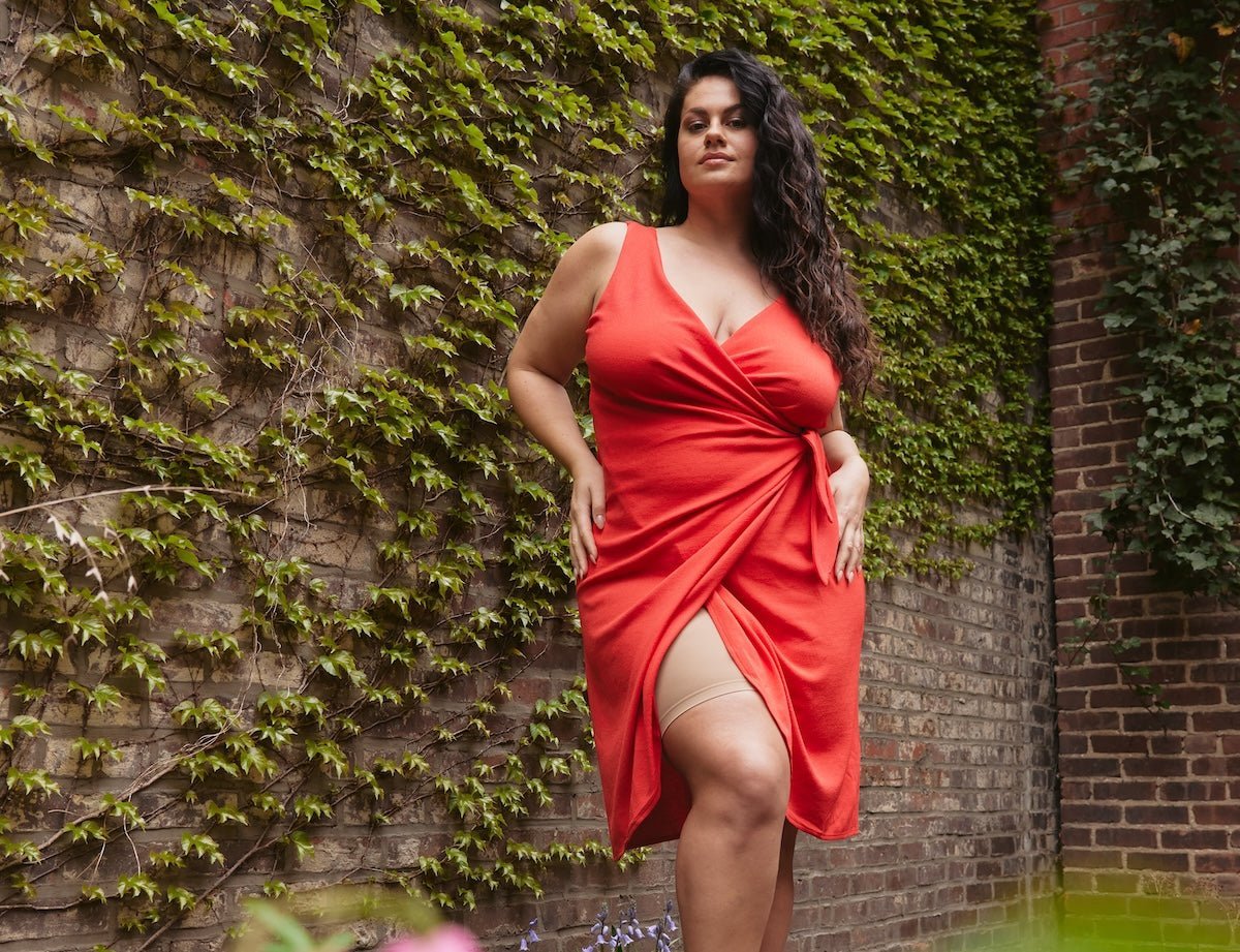 Plus-size model standing in a red wrap dress with a thigh slit showing a beige thigh band against an ivy-covered brick wall
