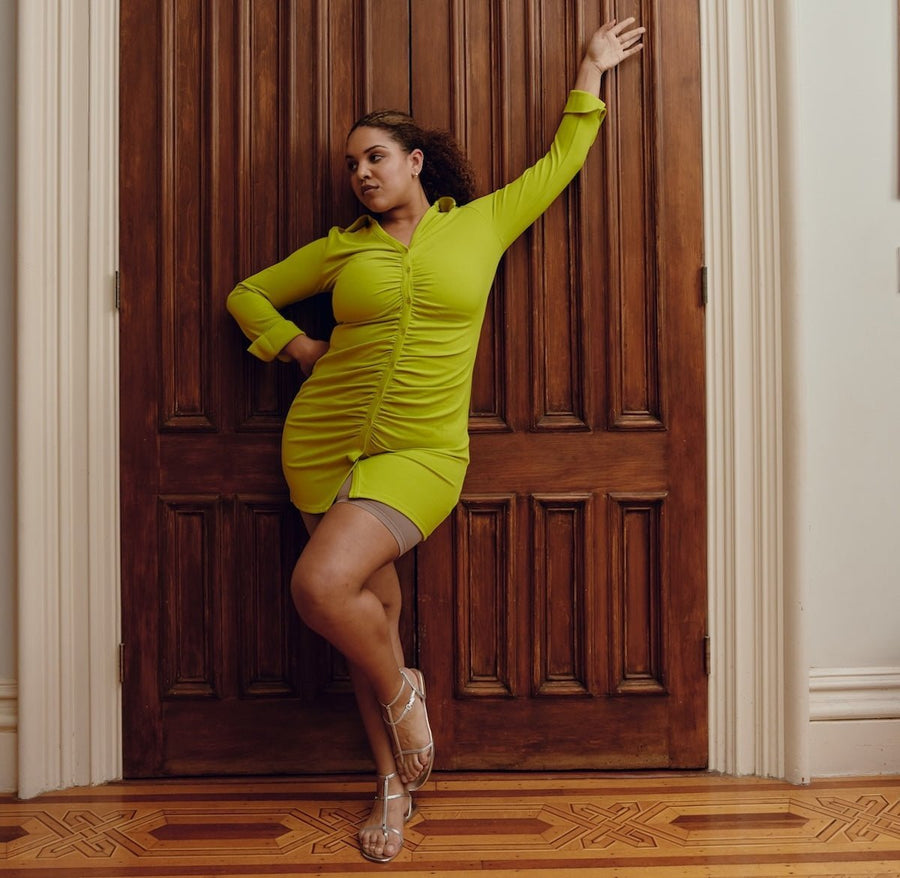 Plus-size model leaning against a wooden door wearing a fitted lime green ruched mini dress and strappy sandals