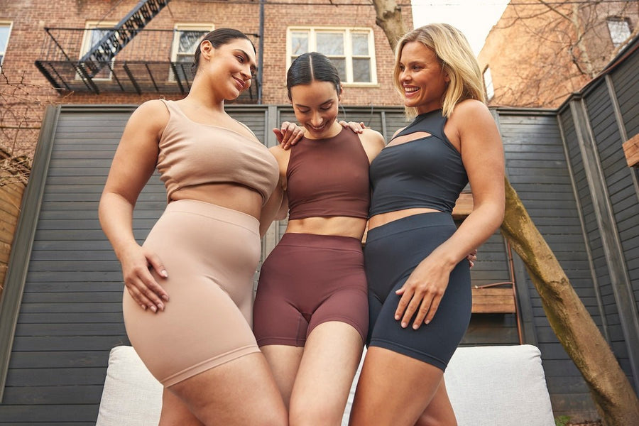 Three women of different body types posing outdoors in nude, brown and black shorties, smiling to show fit on a patio