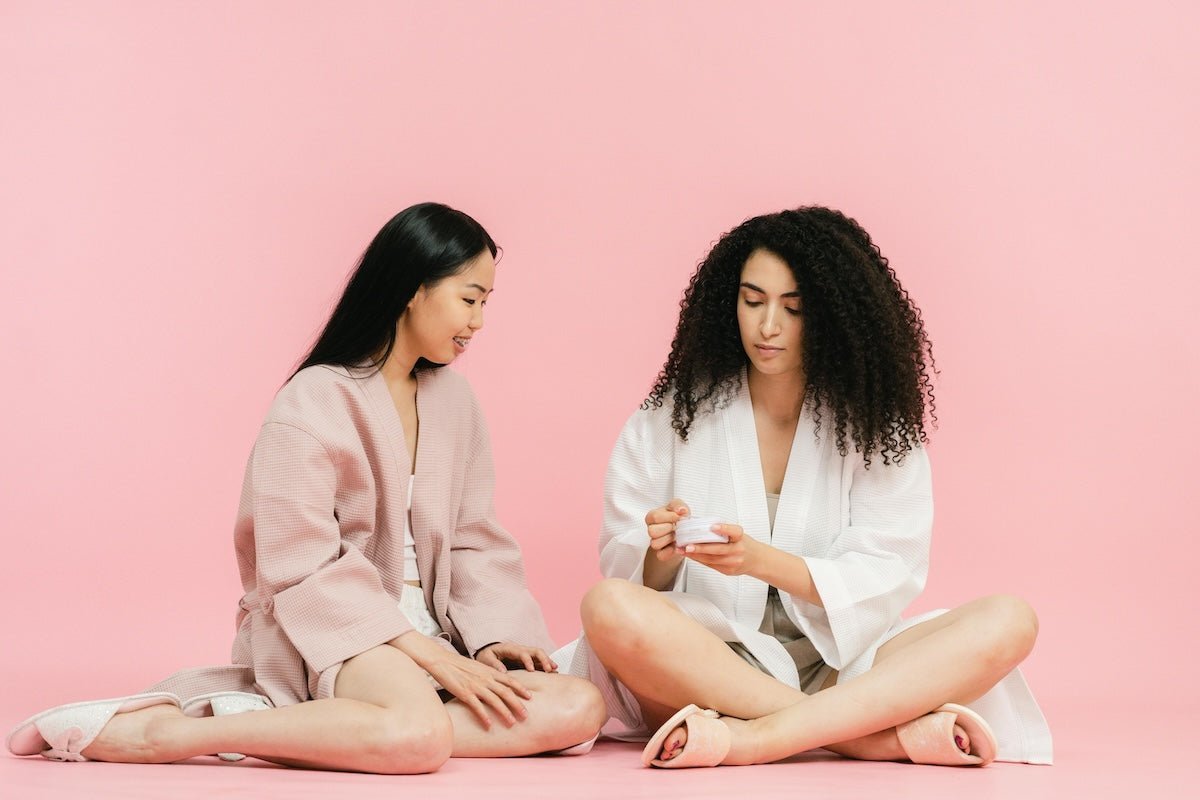 Two women wearing robes sitting on a pink background, one in a white robe opening a jar while the other in a pink robe watches