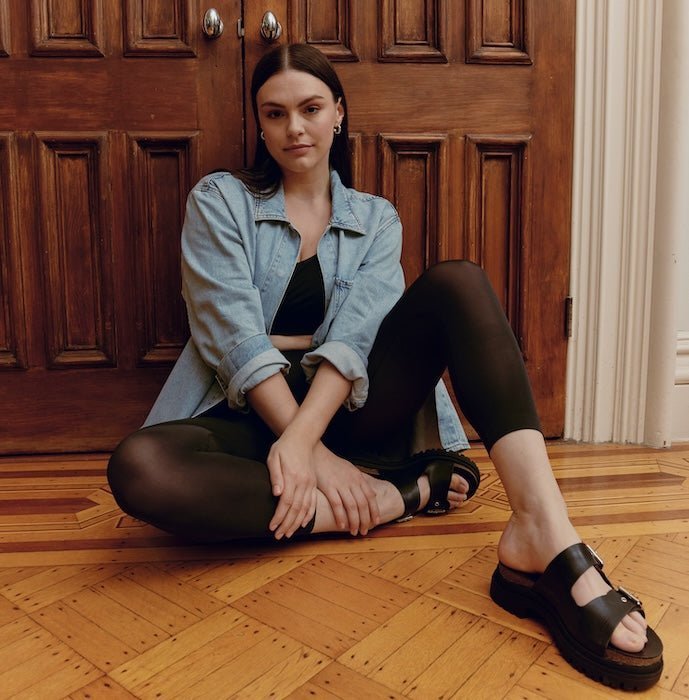 Woman sitting cross-legged wearing black sheer leggings, denim jacket, and black sandals on a parquet floor in front of wooden doors