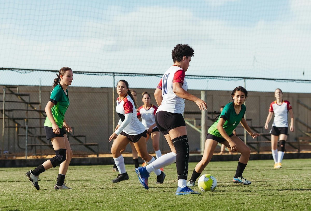 Female soccer players competing for the ball on a sunny outdoor field with bleachers and a chain-link fence in the background