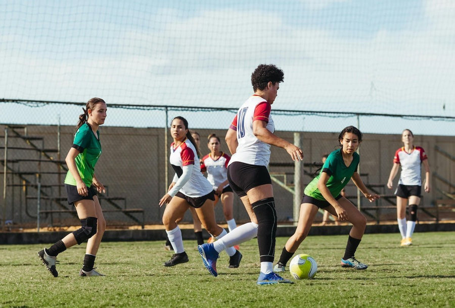 Female soccer players competing for the ball on a sunny outdoor field with bleachers and a chain-link fence in the background