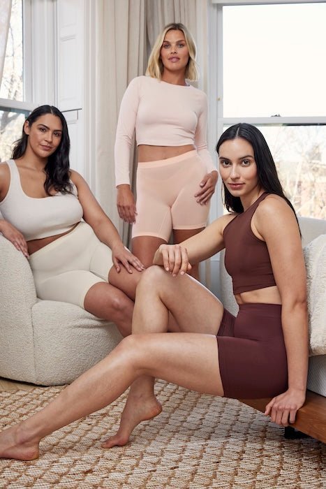 Three women wearing anti-chafing mid-thigh shorts in cream, blush, and chocolate brown posing in a bright living room