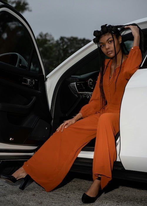 Model sitting in a white car doorway wearing a rust orange long-sleeve jumpsuit with relaxed wide-leg fit, outdoor evening setting