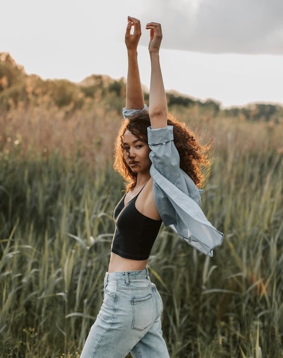 Woman in light-wash baggy jeans and black crop top raising her arms in a grassy field