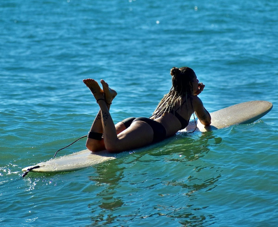 Woman with braided hair lying on a surfboard in a black bikini, resting on calm blue ocean water