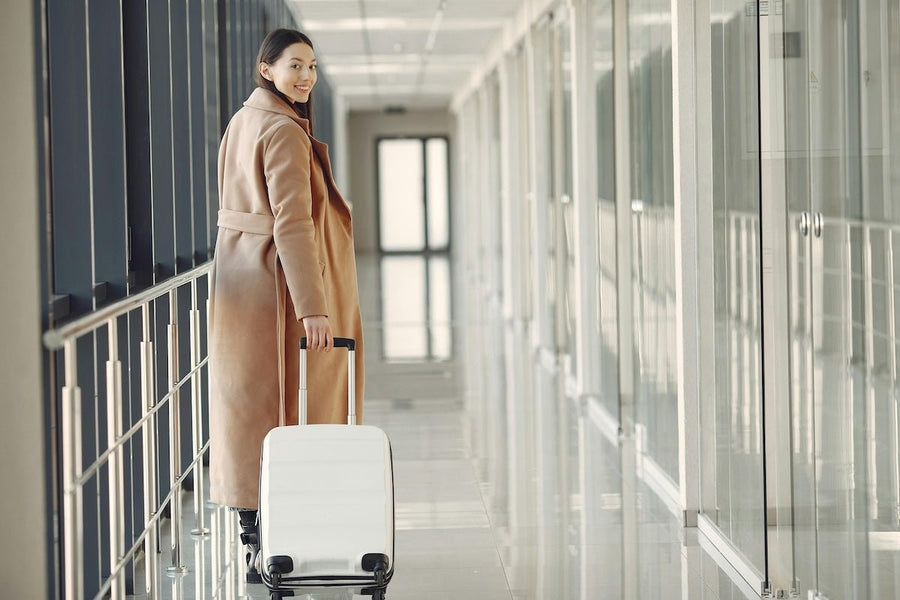 Woman pulling a white carry-on suitcase through a bright glass-sided airport corridor, looking back and smiling