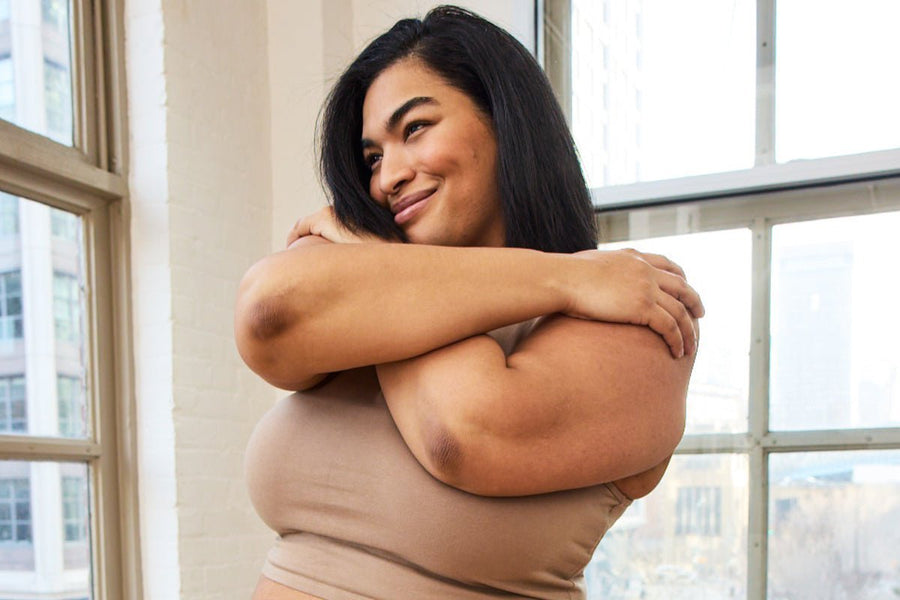 Smiling woman hugging herself wearing a beige fitted tank top in a sunlit loft with large windows