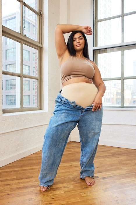Plus-size model posing in a sunlit loft wearing a nude high-waist base layer brief and tan crop bra showing smoothing coverage