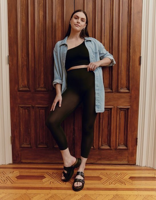 Front view of black high-waist cropped leggings showcasing slim, ankle-length fit on model with denim shirt and wooden door background