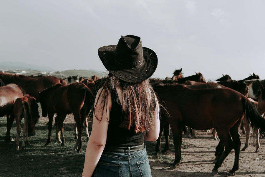 Woman wearing a black cowboy hat, black tank top, and jeans standing with a herd of brown horses in an open field