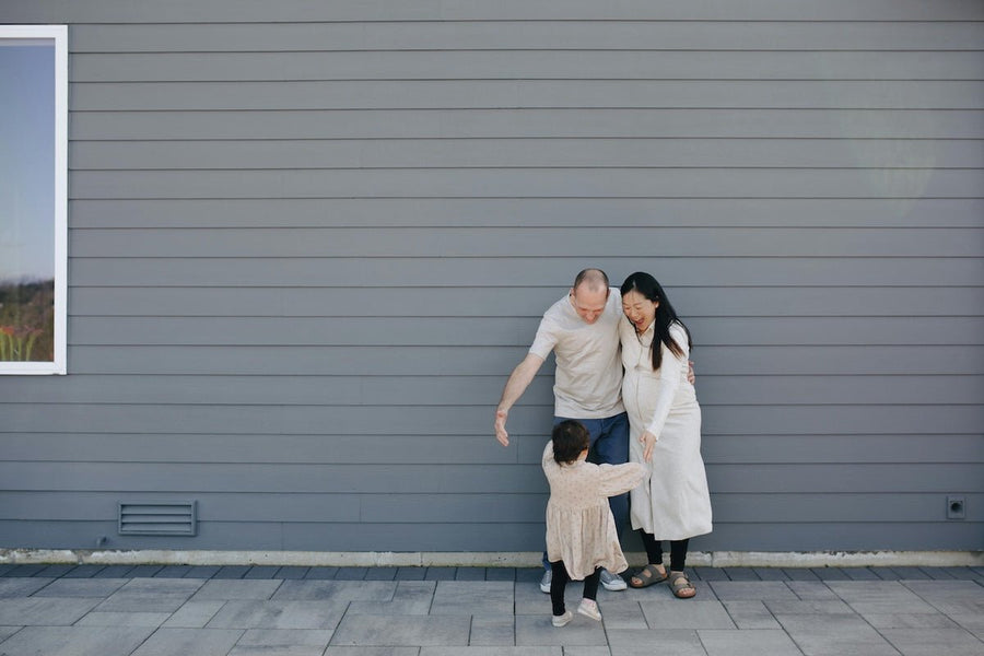 Parents crouching to hug their young child in front of a gray horizontal-siding exterior wall