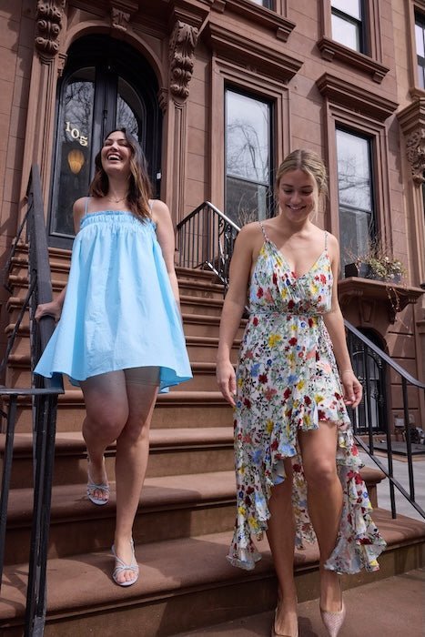 Two women descending brownstone steps, left in a light-blue strapless mini dress, right in a white floral high-low wrap dress