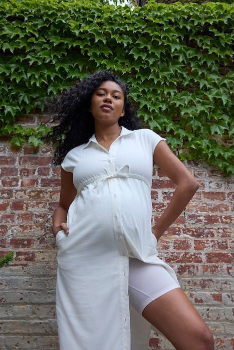 Pregnant woman standing against a brick wall with ivy, wearing a white button-front maternity dress and light beige shorts