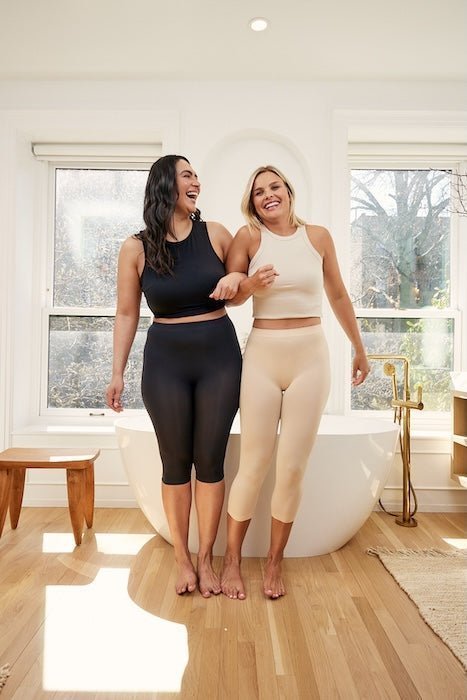 Two women linking arms wearing fitted capri leggings, one black and one beige, in a bright bathroom by a bathtub