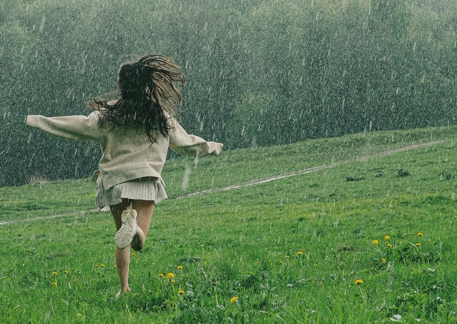 Woman running through rain on a green hillside wearing a cream sweater, pleated skirt, and white sneakers, arms outstretched