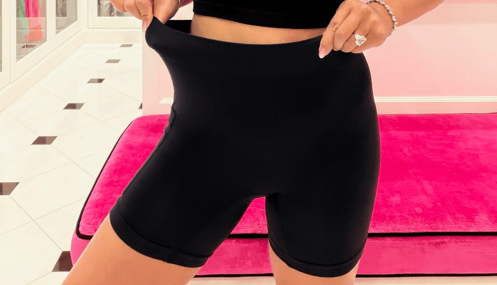 Front close-up of black high-waist shaping shorts highlighting a stretchy waistband and mid-thigh length on a pink bench and tiled floor